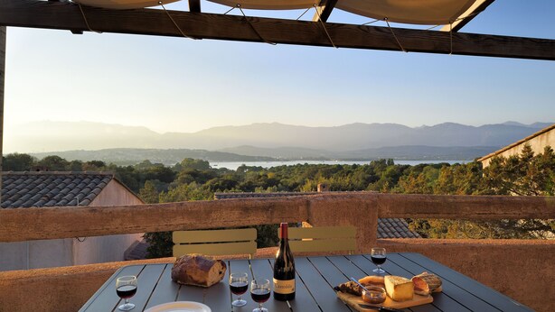 Une table dressée pour un repas, avec une bouteille de vin, des verres, du pain et du fromage, sur un balcon donnant sur un paysage magnifique, avec des montagnes et une étendue d’eau.