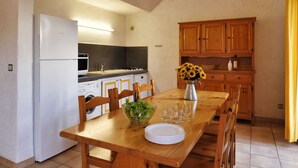 Une cuisine compacte équipée d’un réfrigérateur, d’un four à micro-ondes et d’un lave-linge. On y trouve une table à manger en bois avec des chaises, un saladier rempli de salade et des verres à vin.