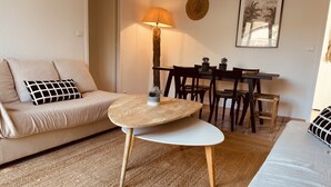 A living room with a beige sofa, a round wooden table, a lamp, and a dining area with chairs and a table. - Beach House (Lège-Cap-Ferret)