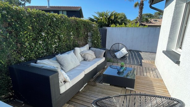 A wooden deck with a sofa and a chair, surrounded by greenery and a white wall. - Beach House (Lège-Cap-Ferret)
