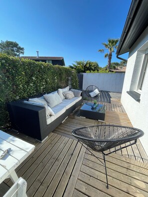 A wooden deck with a sofa and a chair, surrounded by greenery and a white wall. - Beach House (Lège-Cap-Ferret)