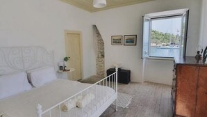 A bedroom with a bed, a dresser, a window with a view of the sea, and a brick wall. - Dolce Vivere by Konnect, Gaios Paxos (Paxos)