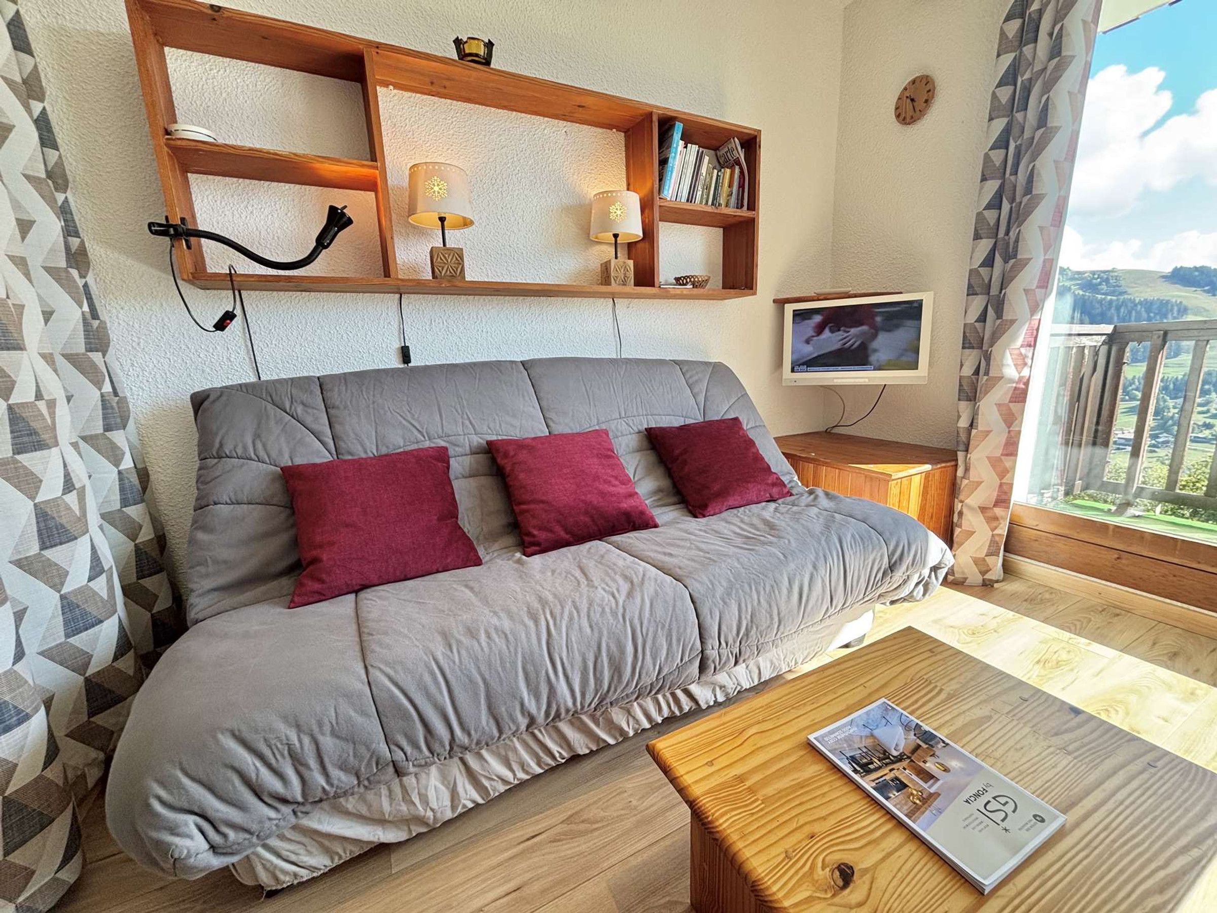 Ein modernes Wohnzimmer mit einem grauen Sofa, roten Kissen, einem Holztisch, einem Bücherregal und einem Fernseher.