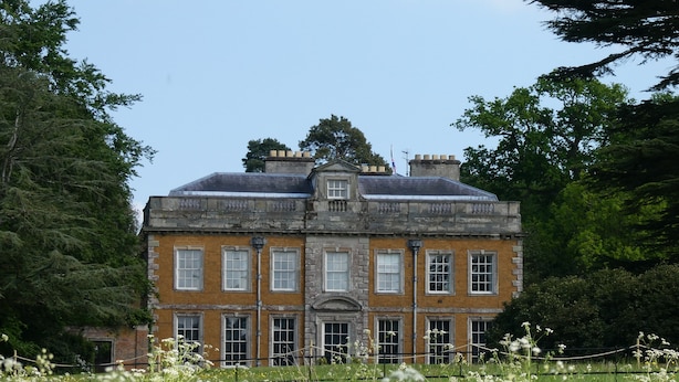 Un grand bâtiment historique, doté de nombreuses fenêtres et d’une entrée imposante, entouré d’une végétation luxuriante et d’un champ de fleurs sauvages.