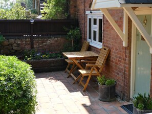 Een terras met een houten tafel en stoelen, een bakstenen muur en een raam met witte luiken.