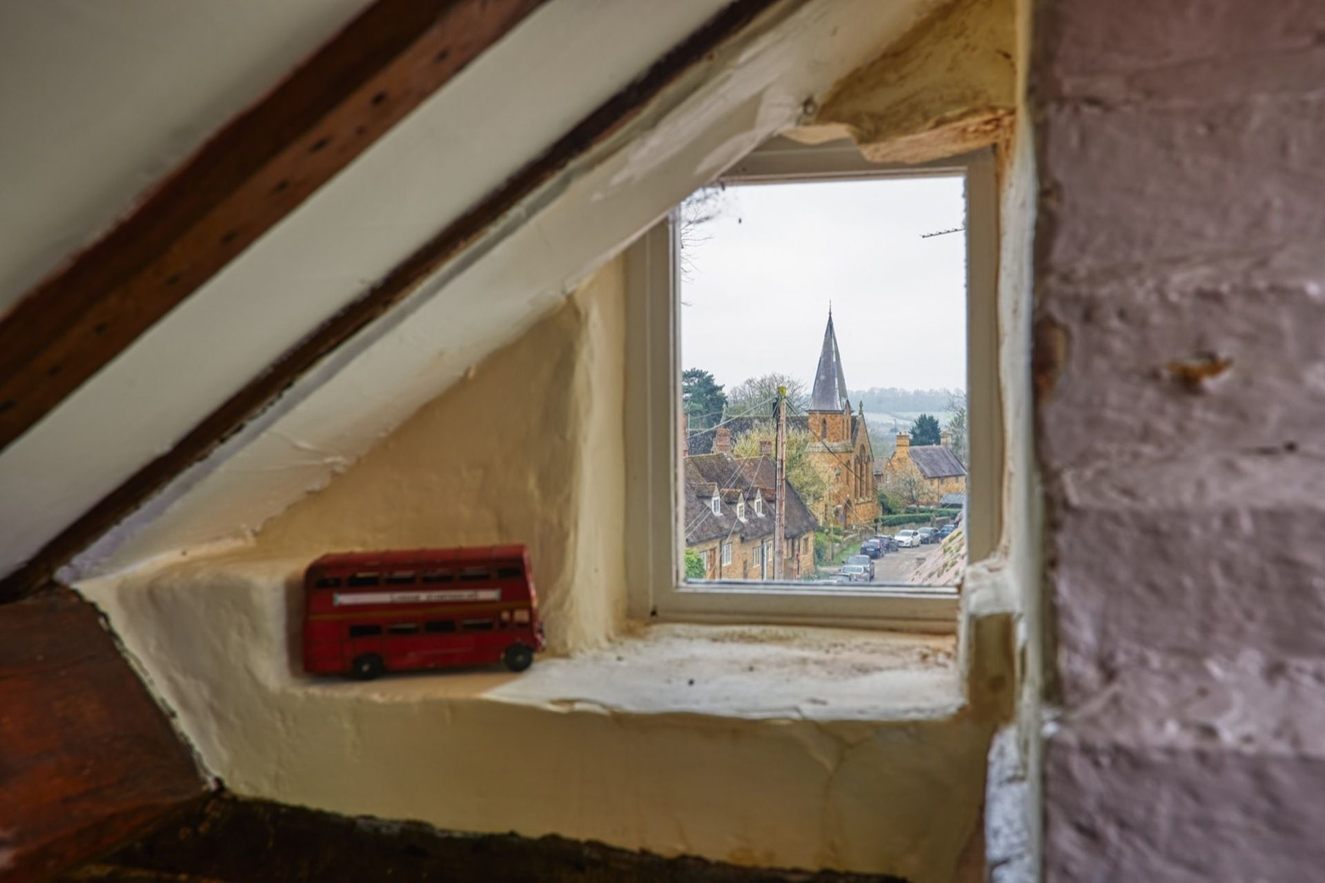 A small attic window with a view of a church and a red bus parked outside.