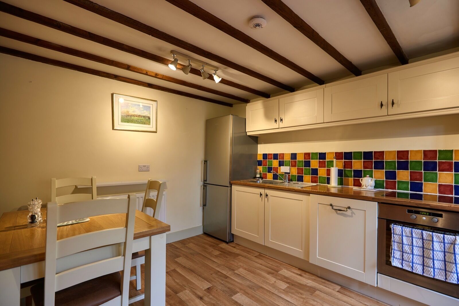 A kitchen with wooden beams, a dining table with chairs, a refrigerator, a colorful backsplash, and a clock.