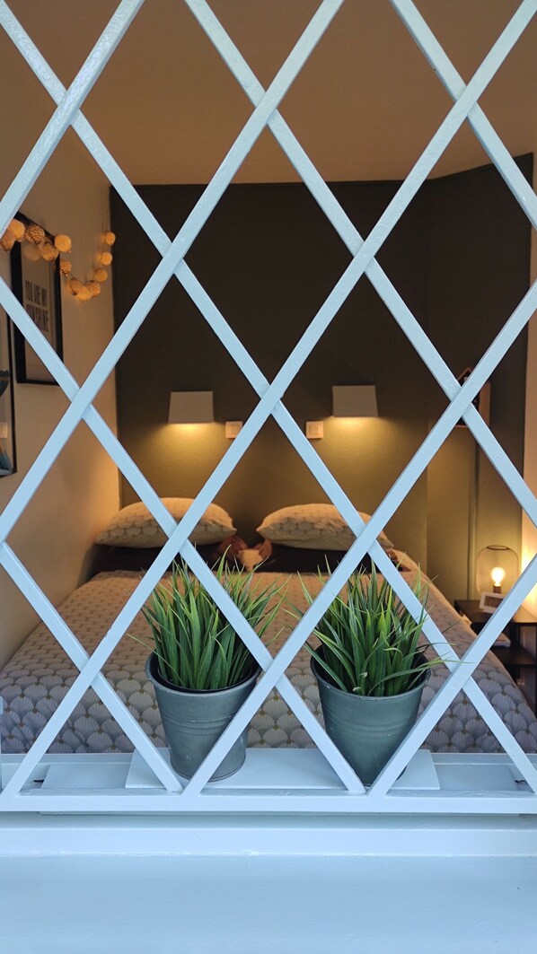 A bedroom with a white lattice partition, two potted plants, a bed with pillows, and a bedside table with a lamp.