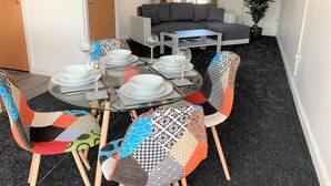 A dining area with a round glass table set for four, surrounded by colorful chairs. A gray sofa is visible in the background. - Toothbrush Apartments - 2 Bed 1 Bath Apt (Ipswich)