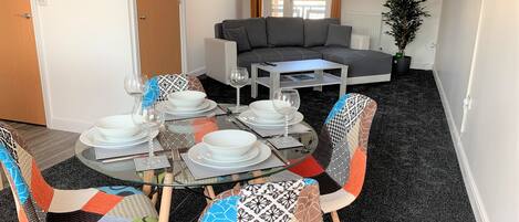A dining area with a round glass table set for four, surrounded by colorful chairs. A gray sofa is visible in the background.