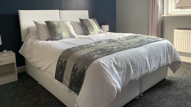 A neatly made bed with white linens and patterned pillows, a dark blue wall, and a window with curtains. - Toothbrush Apartments - 2 Bed 1 Bath Apt (Ipswich)