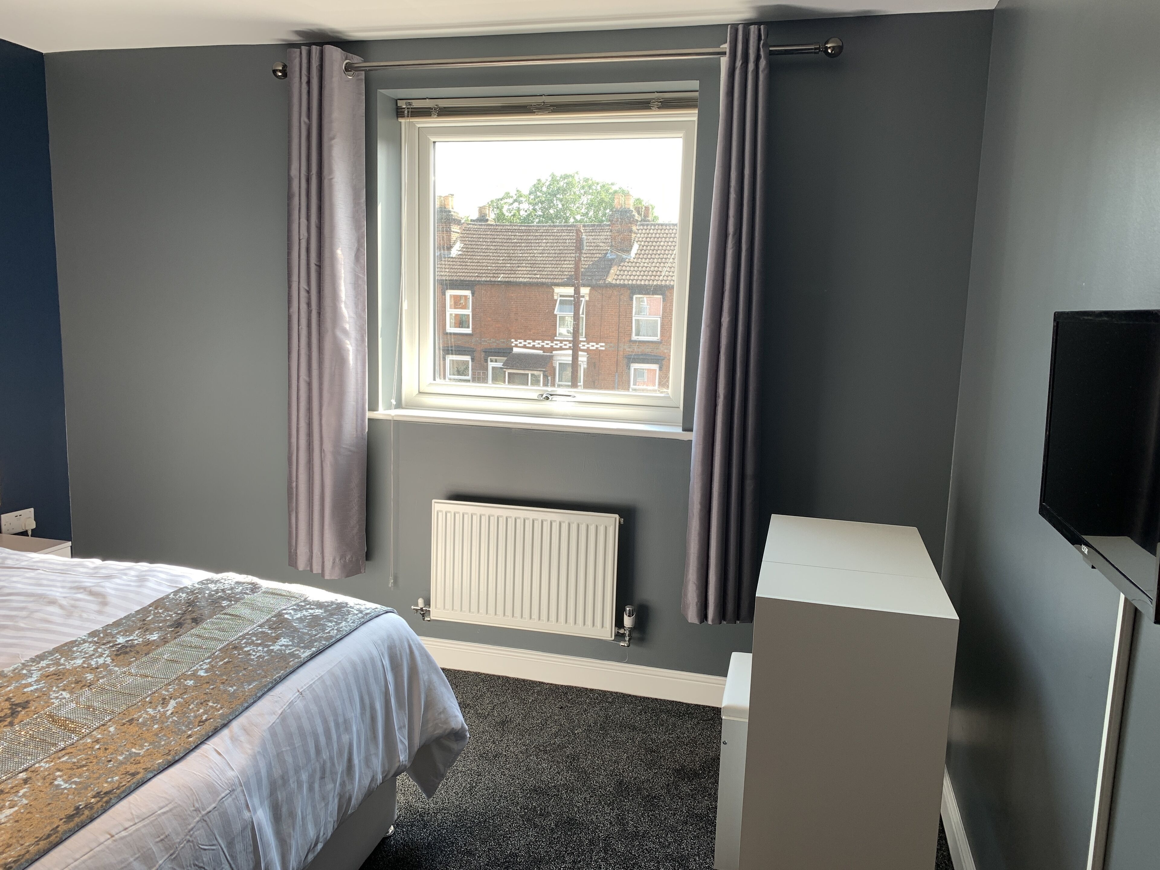 A hotel room with a bed, a radiator, a window with curtains, and a television.