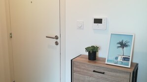 Une pièce avec une porte blanche, un meuble en bois orné d’une plante et d’un tableau, un thermostat et un panneau en bois sur lequel sont inscrits des chiffres et le mot « MAISON ».