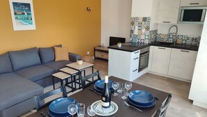 Un salon moderne avec un canapé gris, une table à manger dressée avec de l’assiette bleue et des verres à vin, et une cuisine dotée de meubles blancs et d’un four à micro-ondes.