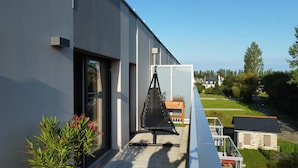 Un bâtiment moderne doté d’un balcon agrémenté d’un fauteuil suspendu, d’une plante en pot et offrant une vue sur une route et la verdure environnante.
