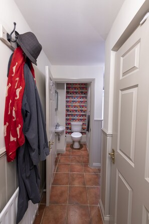 Un couloir étroit, dont le sol est recouvert de carreaux, mène à une salle de bain équipée de toilettes et d’un lavabo.