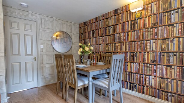 Un coin repas avec une table en bois dressée pour quatre personnes, entourée de chaises, d’un miroir rond et d’une bibliothèque remplie de livres.
