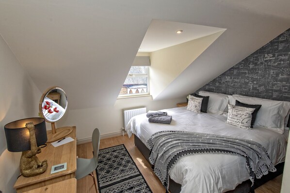Une chambre avec un plafond en pente, un lit avec une literie blanche, un plaid noir et blanc à motifs, un petit bureau avec une lampe et un miroir.