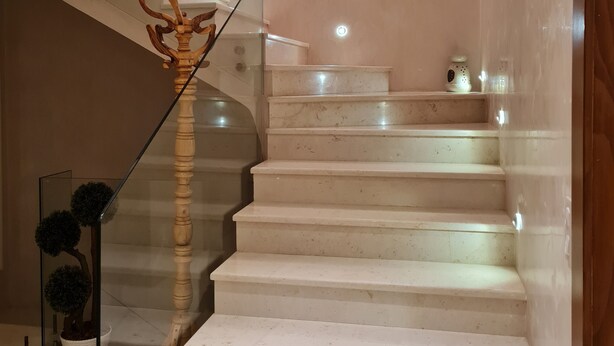 Una escalera con barandas de vidrio y un pasamanos de madera, que conduce a un rellano con una pequeña mesa y objetos decorativos.