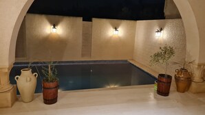Un área de piscina exterior con suelo de baldosas, rodeada de paredes y arcos color beige. Hay tres luces fijadas a la pared y varias plantas en macetas.