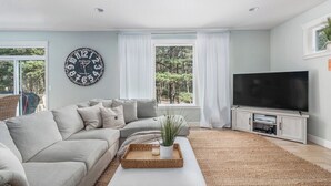A living room with a sectional sofa, a flat-screen TV, a wall clock, and large windows with curtains.