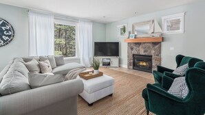 A living room with a sofa, ottoman, and a green armchair. There's a TV, fireplace, and a clock on the wall.