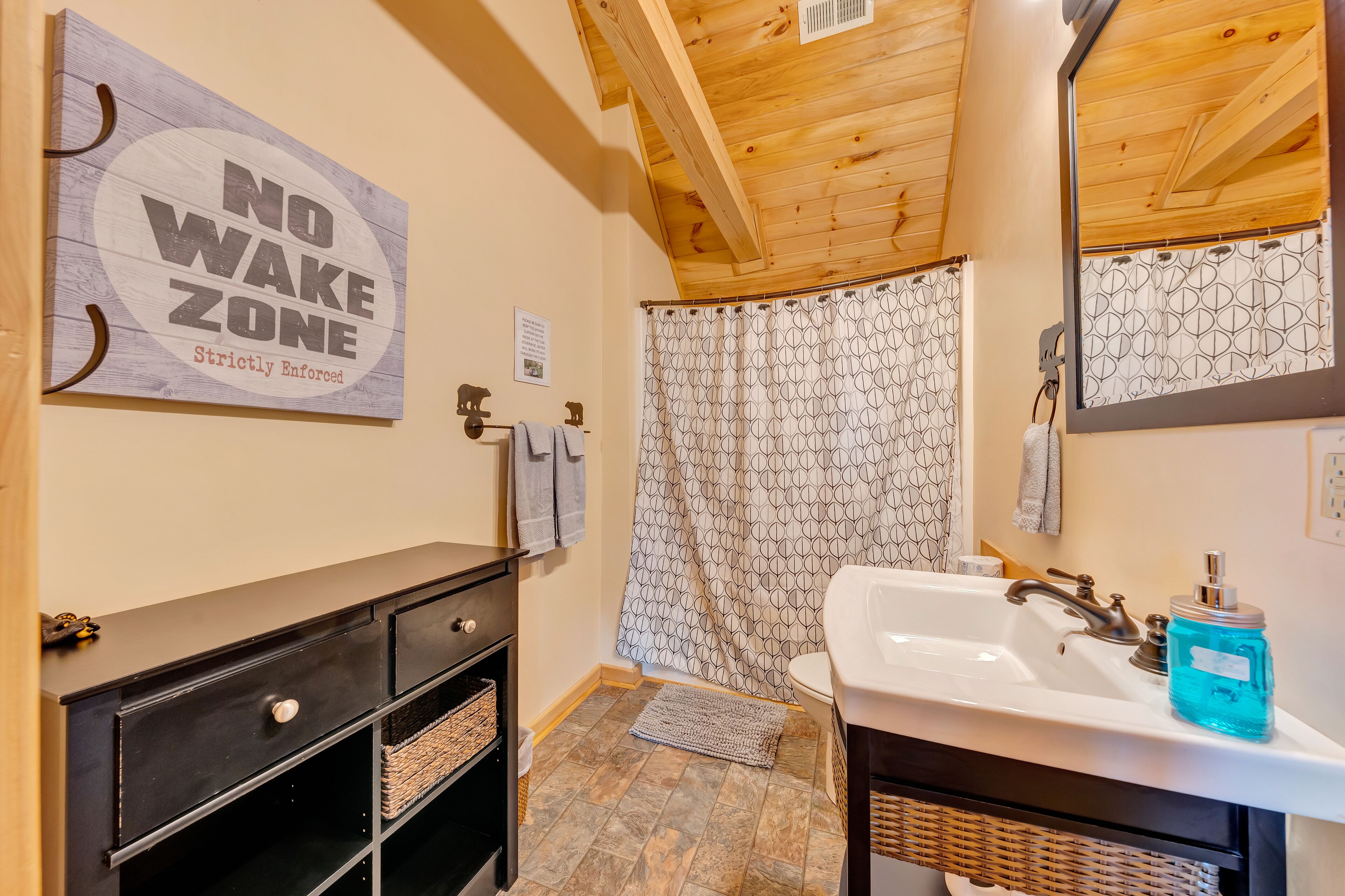 A bathroom with a wooden ceiling, a shower curtain, a sink, and a 'No Wake Zone' sign.