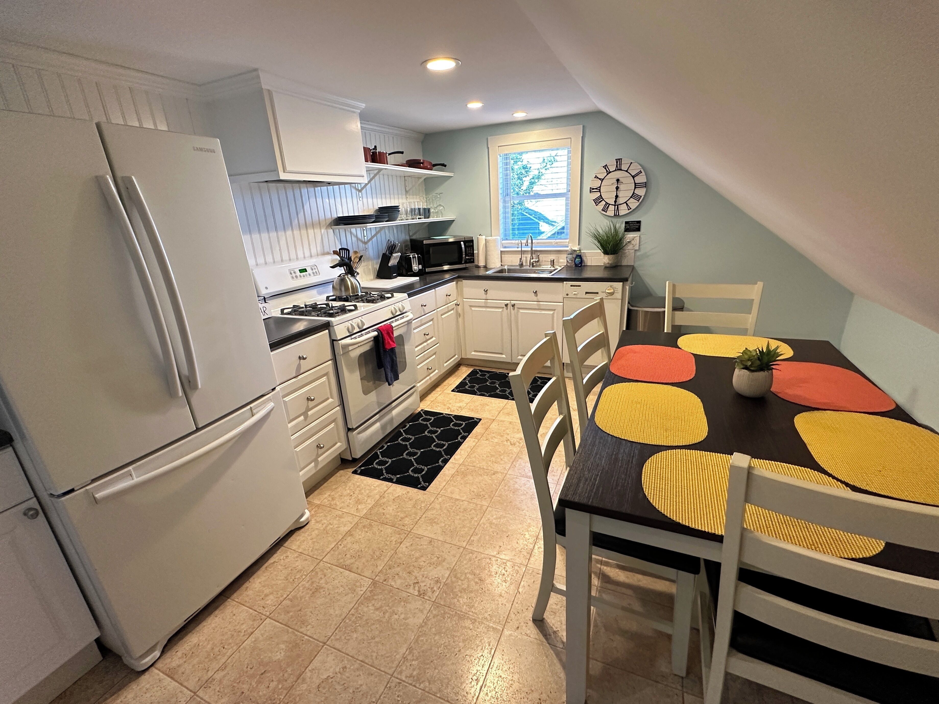 A compact kitchen with white cabinets, a refrigerator, a stove, and a microwave. There is a dining table with yellow and red placemats and a potted plant.