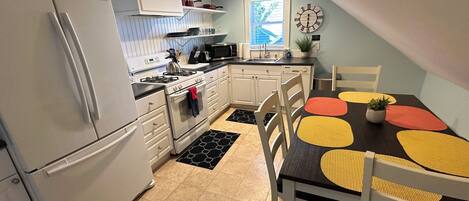 A compact kitchen with white cabinets, a refrigerator, a stove, and a microwave. There is a dining table with yellow and red placemats and a potted plant.