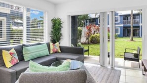 Ein modernes Wohnzimmer mit einem grauen Sofa, bunten Kissen, einer Glastür, die in einen Garten führt, und Blick auf ein blaues Gebäude.