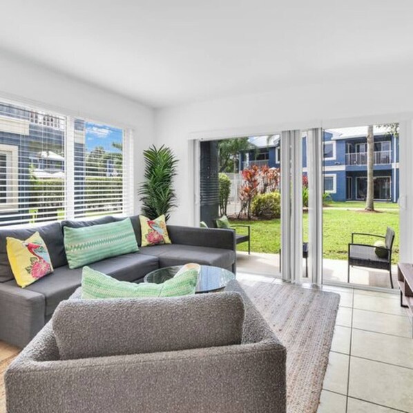 Een moderne woonkamer met een grijze bank, kleurrijke kussens, een glazen deur naar een tuin en uitzicht op een blauw gebouw.