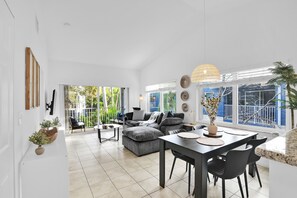 Un vaste espace de vie comprenant un grand canapé d’angle gris, une table à manger avec des chaises et un balcon offrant une vue sur la verdure.