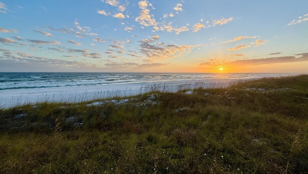 Paisagem costeira com um pôr do sol sobre o oceano, dunas gramadas e céu claro.