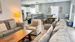 Uma sala de estar moderna com um sofá, poltrona e mesa de centro. Uma área de jantar com mesa e cadeiras é visível ao fundo.