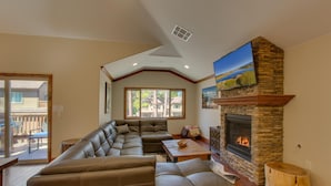 A spacious living room with a large L-shaped sectional sofa, a stone fireplace, and a large window providing a view of the outdoors. - AC, Custom Bunks, Bubble Hockey, Sauna, Walk to Heavenly/ Lake, Grill – Tahoe Woods Paradise (South Lake Tahoe)