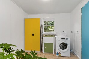 Ein Waschraum mit gelber Tür, ein grüner Hauswirtschaftsraum, ein Fenster mit Blick auf Grünflächen und eine weiße Waschmaschine.