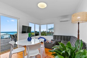 A modern living room with a view of the ocean, a round table with chairs, a grey sofa, and a large plant.