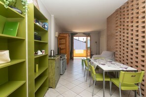 A room with a dining table, chairs, a green bookshelf, a kitchen area, and a brick wall.