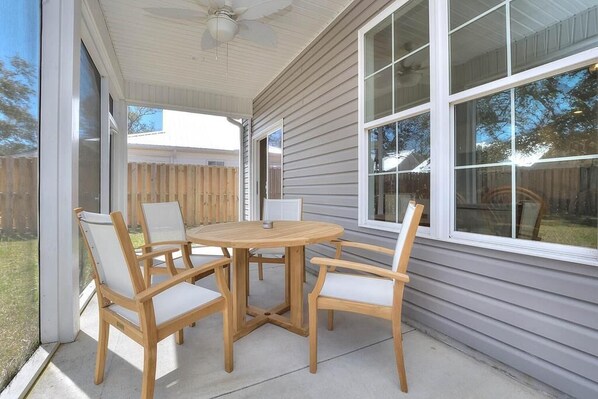 Une terrasse couverte avec une table ronde en bois et quatre chaises, de grandes fenêtres et un ventilateur de plafond.