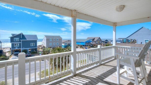 A covered porch with a white railing overlooking a beach with several houses.