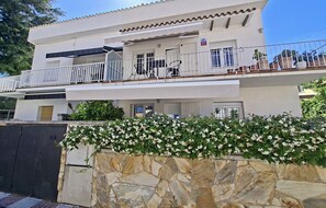 A two-story building with balconies, a stone wall with flowering plants, and a clear sky.