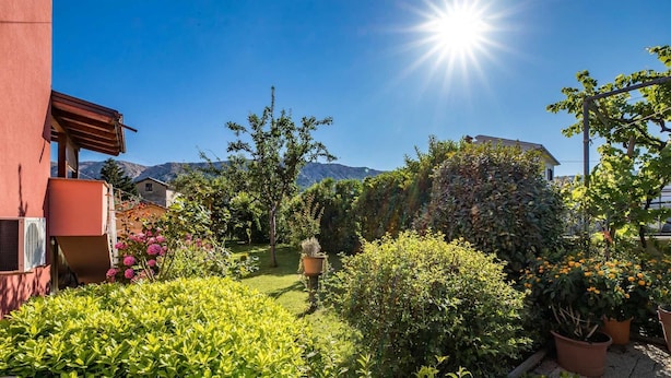 A garden with various plants and flowers, a building with a red facade, and mountains in the background. - Nadica Souterrain - Cozy 1BR Apartment for Two (Baška)