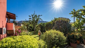 Un giardino con diverse piante e fiori, un edificio con facciata rossa e montagne sullo sfondo.