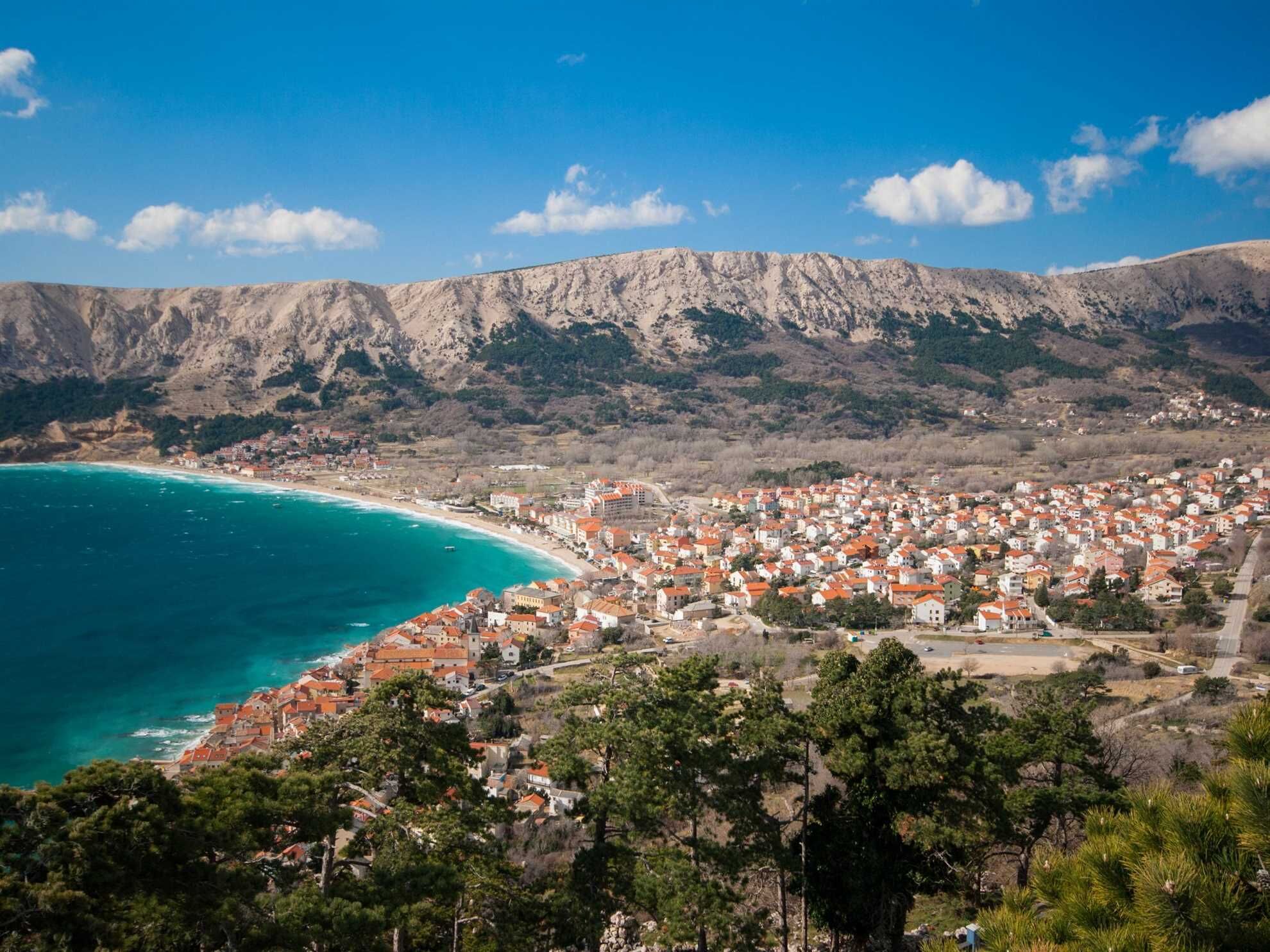 A coastal town with buildings along the shoreline, a mountain range in the background, and a clear sky.