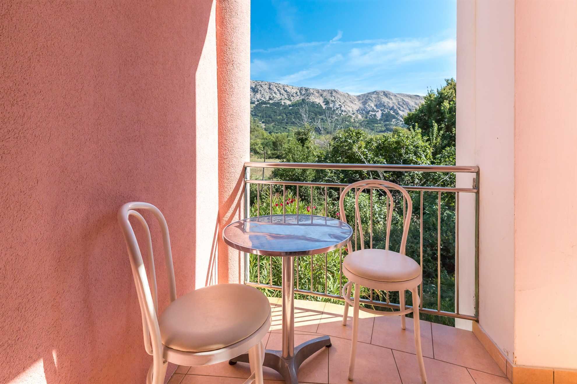 En balkon med to stole og et bord, der giver udsigt til bjerge og grønne omgivelser.