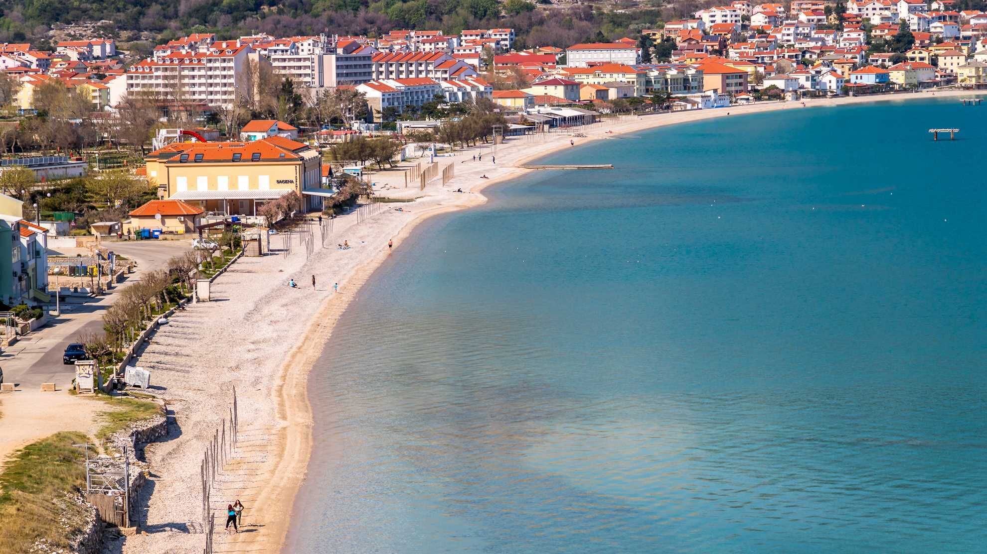 Une ville côtière dotée d’une plage de sable, de bâtiments aux toits rouges et d’une mer d’un bleu limpide.