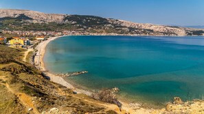 A coastal town with a clear blue sea, a sandy beach, and a mountainous backdrop.