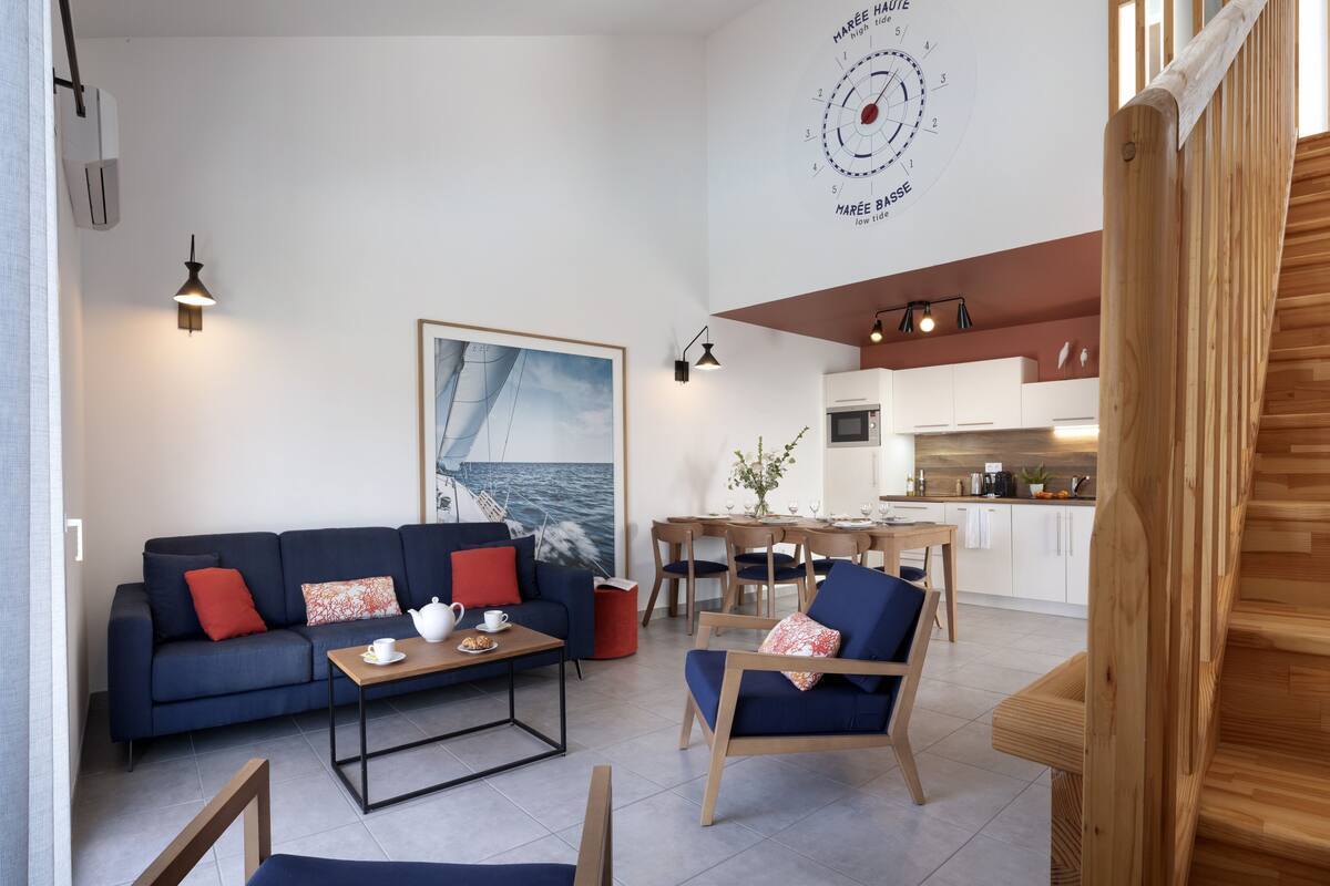 Una sala de estar moderna con sofá, mesa de centro y sillas, un comedor con mesa y sillas, una cocina con armarios y electrodomésticos, y una escalera de madera.