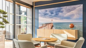 Una habitación moderna con un ventanal grande, dos sillones grises, una mesa de madera y un cuadro de un faro en un acantilado.