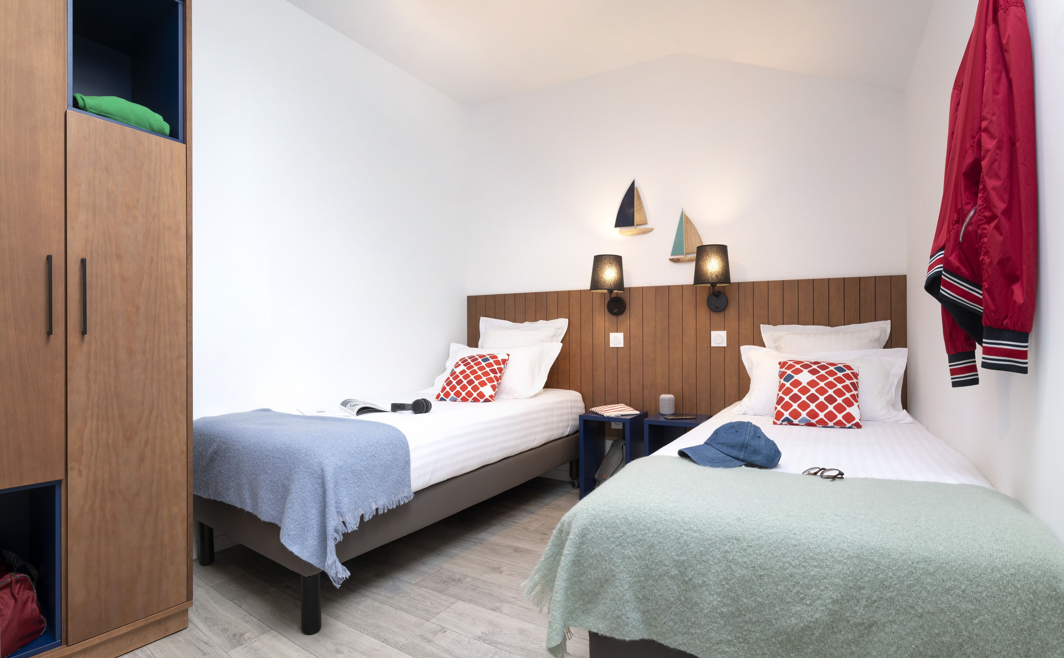 A hotel room with two single beds, wooden wardrobe, and a nightstand with a lamp.